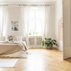 Sypialnia z łóżkiem w kolorze cappuccino, idealnie komponującym się z jasnymi zasłonami, drewnianą podłogą i zieloną rośliną.