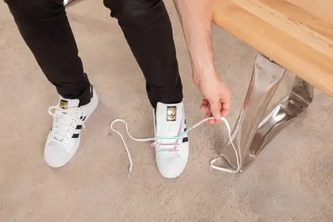 Instrukcja jak wiązać sneakersy: białe buty Adidas z czarnymi paskami, osoba zawiązuje sznurówki.