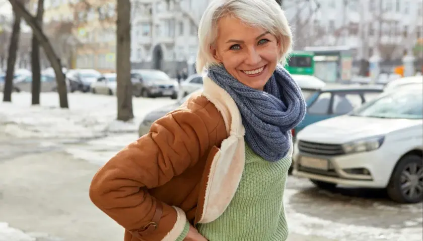 Uśmiechnięta kobieta w brązowym kożuszku zimowym, z niebieskim szalikiem i zielonym swetrem, pozuje na tle zimowego miasta.