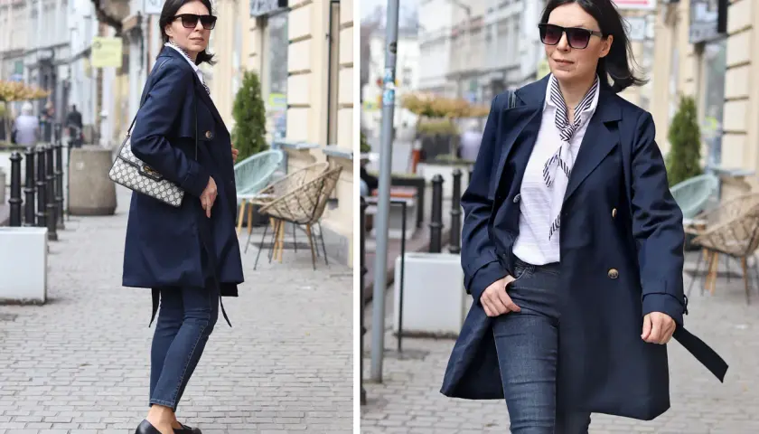 Elegancka kobieta w granatowym trenczu damskim, białej bluzce w paski i jeansach spaceruje po brukowanej ulicy.
